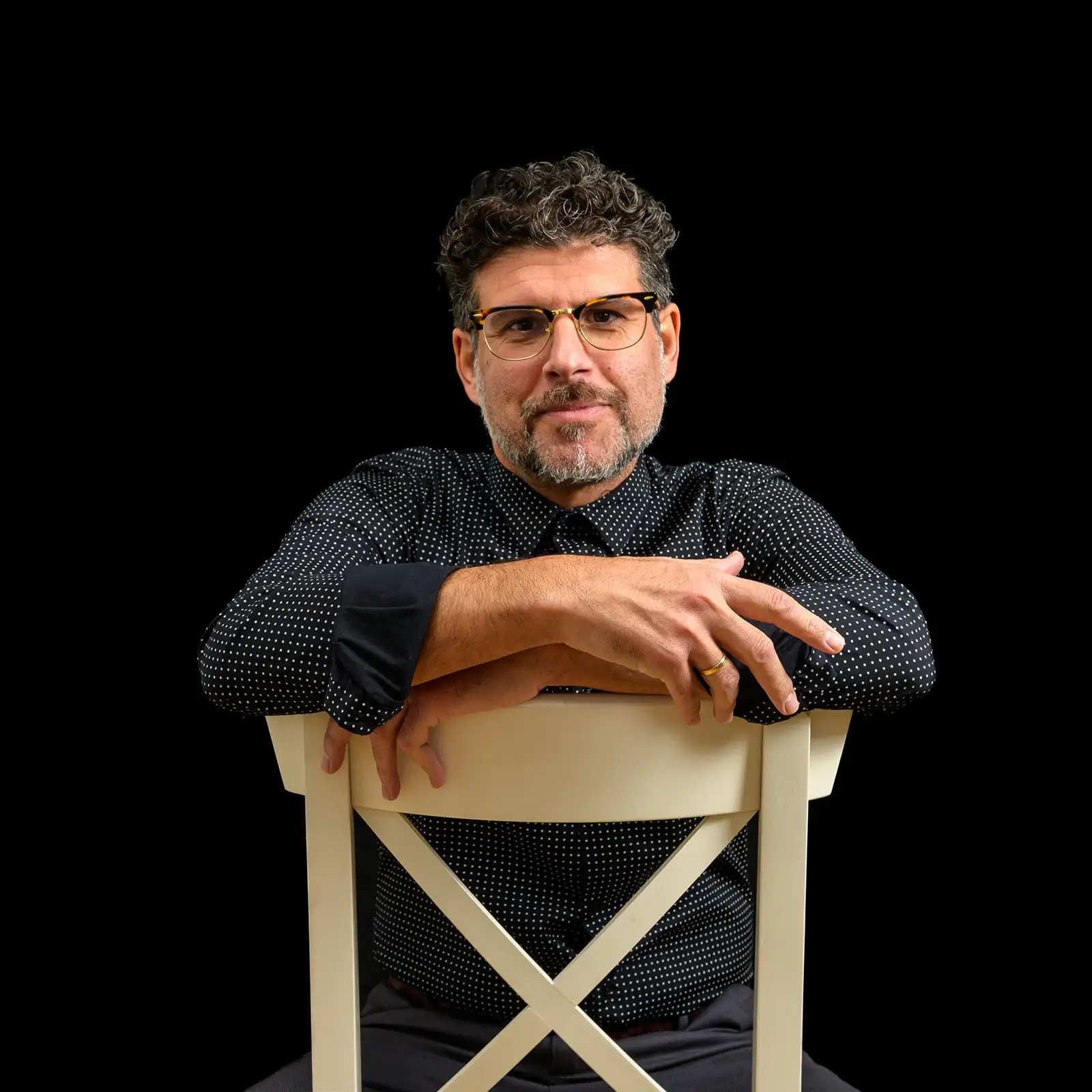 martinez-picon-Retrato Retrato del Dr. Jose María Martínez Picón, psicólogo, sentado con los brazos cruzados sobre el respaldo de una silla, sobre fondo negro